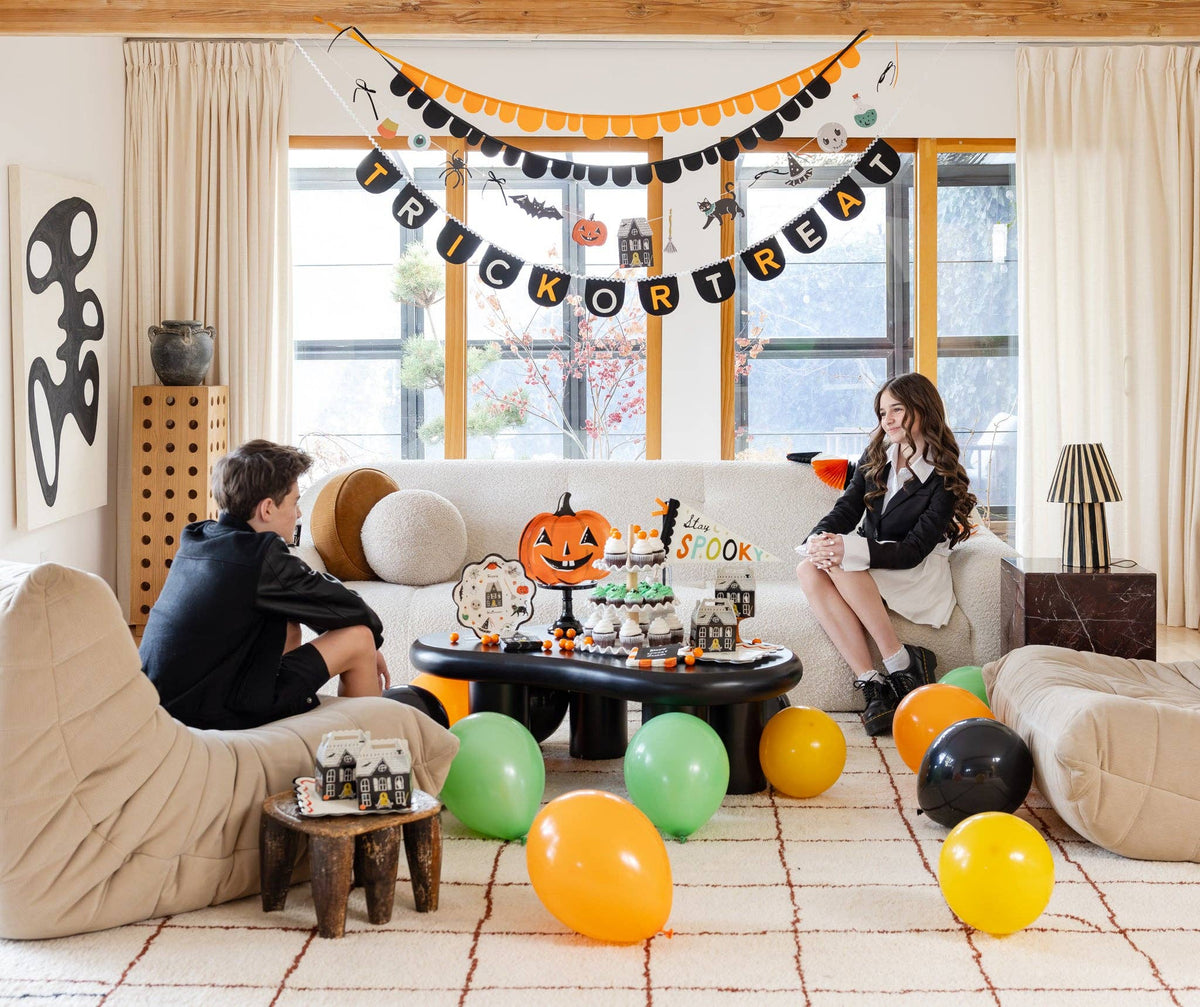 Wide shot of a living room decorated for a kids' Halloween party. The illustrated paper character garland hangs across a window at mid-height, below a layered display of the "Trick or Treat" felt banner (bottom) and an orange-and-black scalloped paper garland (top). Die-cut characters (spider, bat, pumpkin, haunted house, cat, skull, potion bottle) are clearly visible across the garland's span. Two children in black sit on a white sofa and beige chair. 