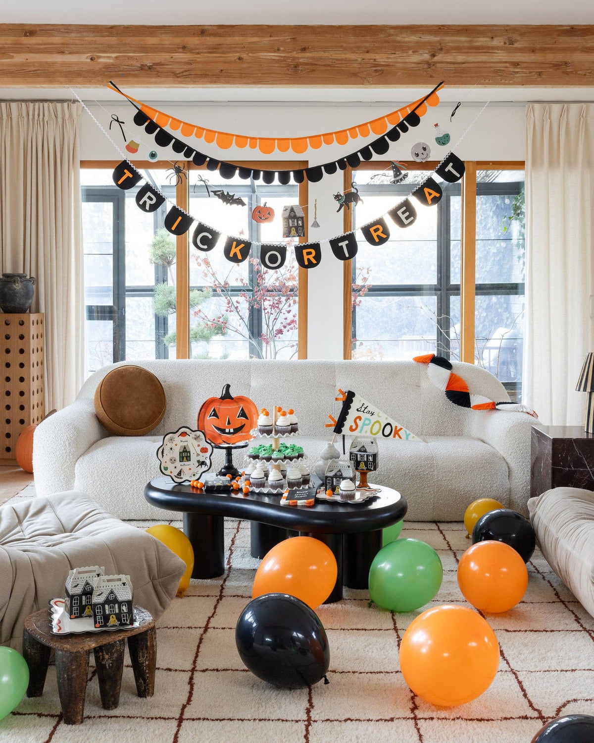 Same room and setup as Image 1 without children visible. Three-layer garland display clearly visible: orange-and-black scalloped garland on top, the character paper banner in the middle, and the Trick or Treat felt banner at the bottom. Dessert table, balloons, haunted house favor boxes, and pumpkin plate visible below.