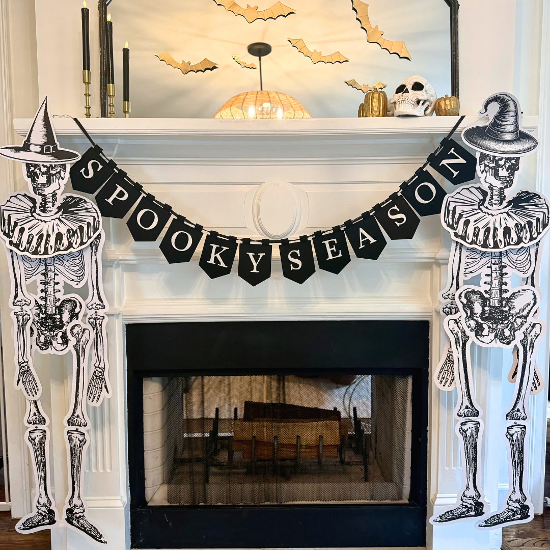 White fireplace mantel decorated for Halloween with a black "Spooky Season" pennant banner, jointed paper skeletons in witch hats on each side, gold paper bats on the mirror, a skull, gold pumpkins, and black taper candles.