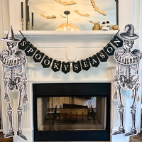 White fireplace mantel decorated for Halloween with a black "Spooky Season" pennant banner, jointed paper skeletons in witch hats on each side, gold paper bats on the mirror, a skull, gold pumpkins, and black taper candles.