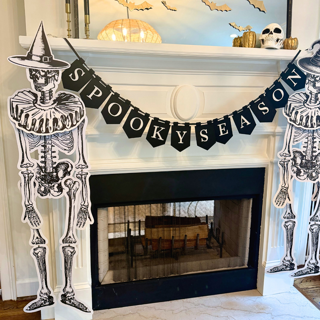 Angled view of a Halloween fireplace with two jointed paper skeletons in witch hats framing a black "Spooky Season" banner, gold bats and a skull on the mantel, and a marble hearth below.