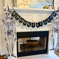 Angled view of a Halloween fireplace with two jointed paper skeletons in witch hats framing a black "Spooky Season" banner, gold bats and a skull on the mantel, and a marble hearth below.
