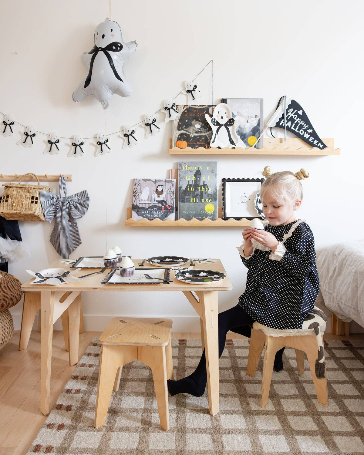 Child sitting at a small wooden table with Halloween decorations on shelves above, and ghost-shaped napkins arranged on a dessert table at a spooky soirée.