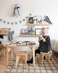 Child sitting at a small wooden table with Halloween decorations on shelves above, and ghost-shaped napkins arranged on a dessert table at a spooky soirée.