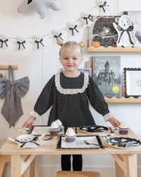 Child in a black polka dot dress standing at a table with Halloween-themed decorations showing the Spooky things ahead paper plates. 