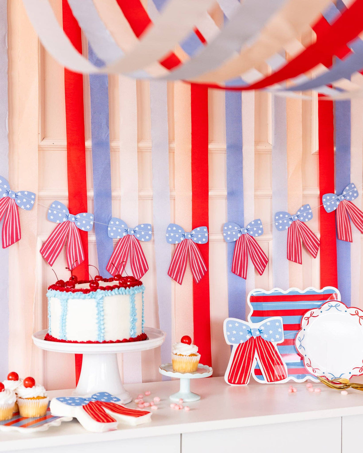 Close-up of patriotic dessert table with stars and stripes bow banner hanging over cake stand, bow-shaped plates, and striped paper plates