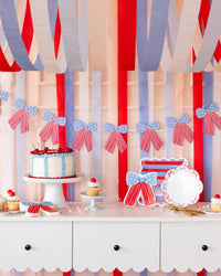 Full dessert table styled with stars and stripes bow banner, red white and blue cake, cupcakes, bow-shaped paper plates, and crepe streamers