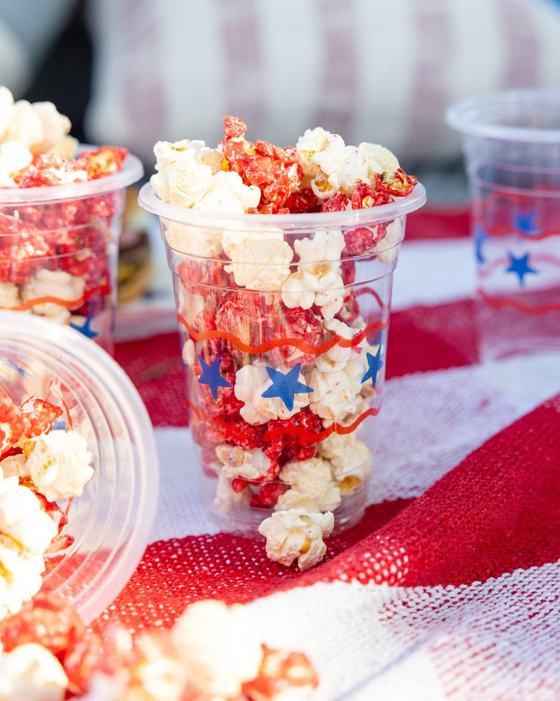 Stars and stripes clear plastic party cups filled with red and white popcorn on a red and white striped tablecloth