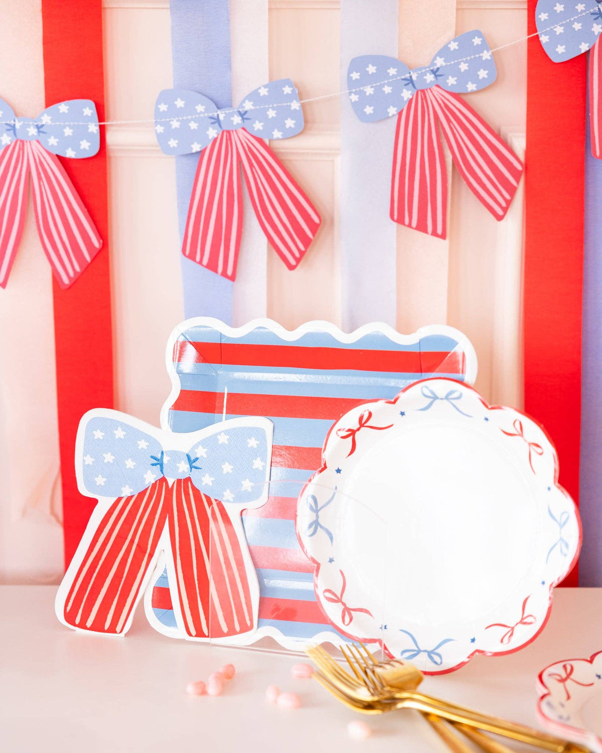 Stars and stripes collection display showing bow shaped napkin, red and blue striped scalloped plate, and bow scalloped plate with bow banner and gold forks