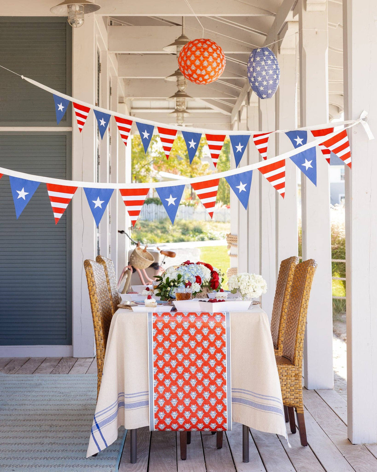 Close-up of stars and stripes pennant banner draped across porch with patriotic table setting, hydrangeas, and printed paper lanterns visible below