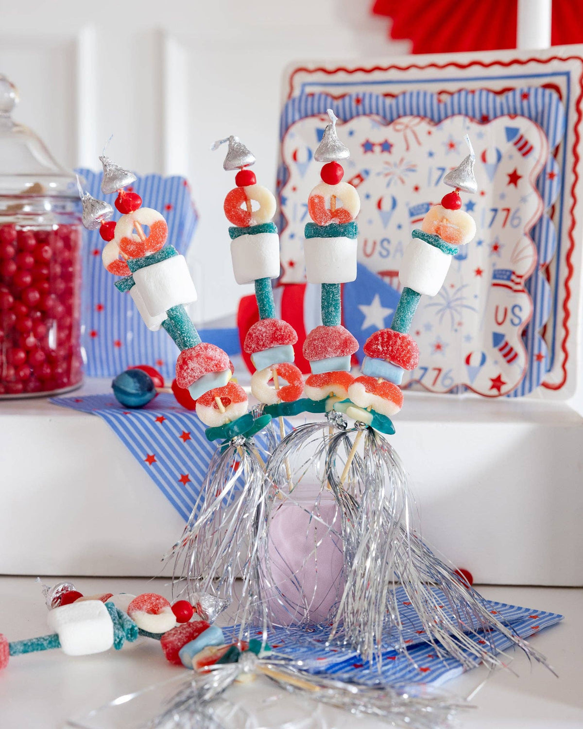 Styled Fourth of July party scene featuring stars and stripes guest napkins beneath candy kabobs with coordinating Americana plates and decor in background