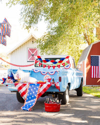 Wide shot of a patriotic truck bed party with stars and stripes paper table runner draped over the tailgate alongside coordinating plates, banners, and a red drink bucket