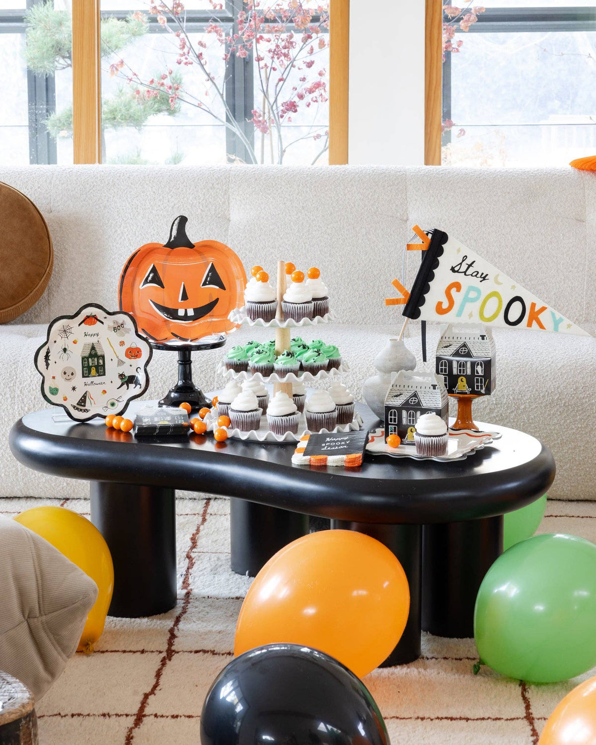 Styled Halloween dessert table on a black coffee table: a jack-o-lantern plate on a black pedestal, a Happy Halloween scalloped plate, a tiered cupcake stand, haunted house treat boxes, and the "Stay Spooky" felt pennant standing upright in a stone vase at right; orange, green, yellow, and black balloons on the floor and autumn branches in the window. Wide lifestyle shot.