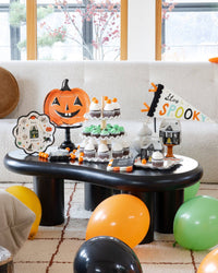 Styled Halloween dessert table on a black coffee table: a jack-o-lantern plate on a black pedestal, a Happy Halloween scalloped plate, a tiered cupcake stand, haunted house treat boxes, and the "Stay Spooky" felt pennant standing upright in a stone vase at right; orange, green, yellow, and black balloons on the floor and autumn branches in the window. Wide lifestyle shot.
