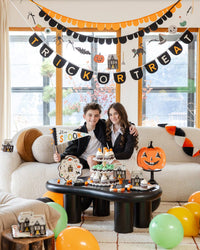 Two smiling teens on a boucle sofa beneath a "TRICK OR TREAT" letter banner and scalloped garland; the boy holds the "Stay Spooky" felt pennant flag by its wood dowel, with a cupcake-and-treat-box dessert table in front and balloons scattered below. Lifestyle portrait.