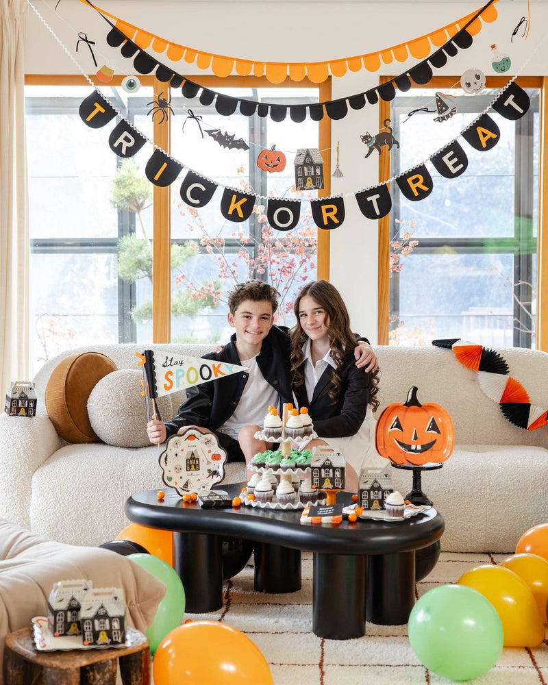 Two smiling teens on a boucle sofa beneath a "TRICK OR TREAT" letter banner and scalloped garland; the boy holds the "Stay Spooky" felt pennant flag by its wood dowel, with a cupcake-and-treat-box dessert table in front and balloons scattered below. Lifestyle portrait.