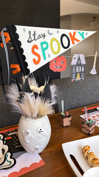 Stay Spooky felt pennant in a white vase with pampas grass on a Halloween table with icon plates and small bites