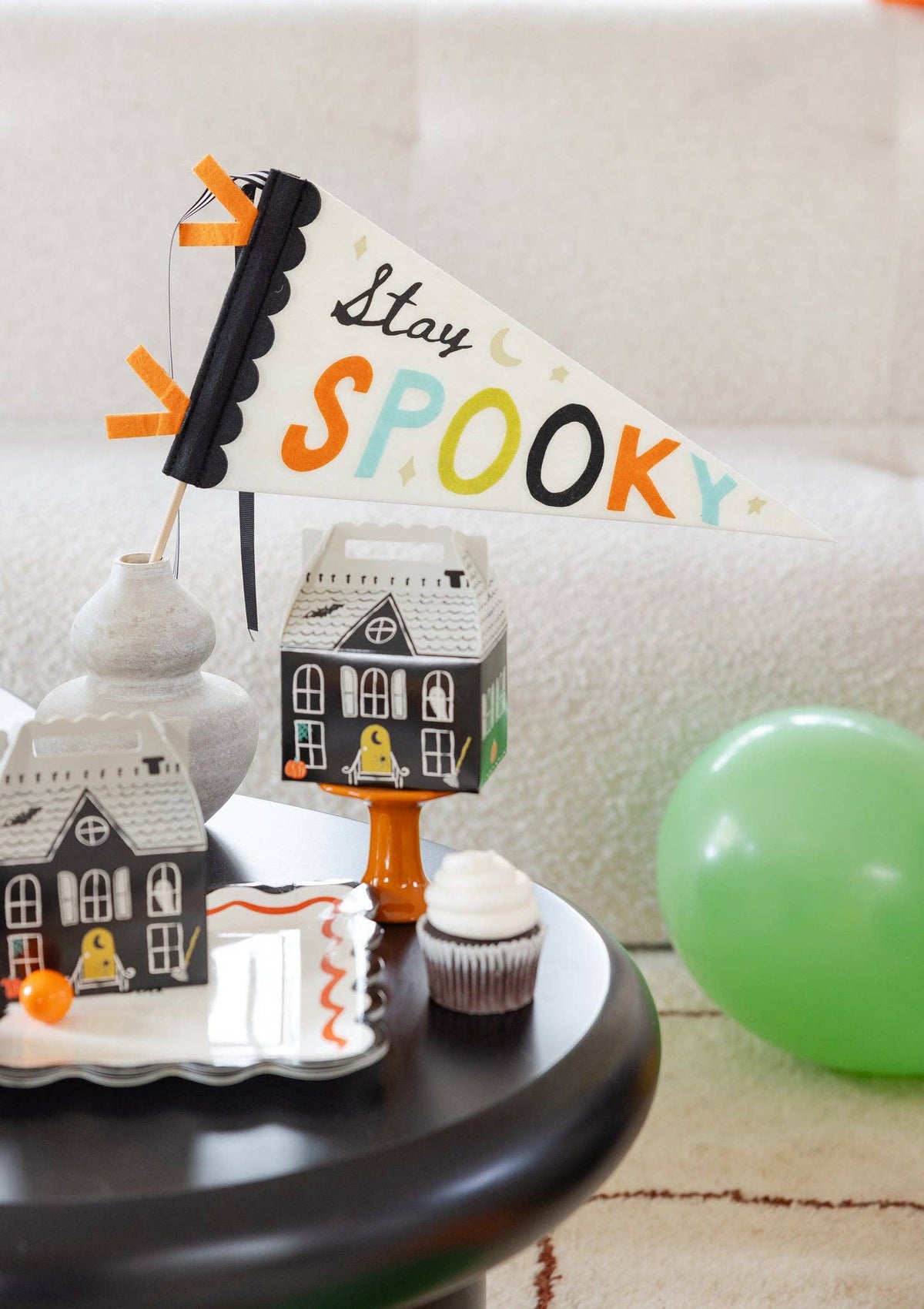 Close-up of the "Stay Spooky" pennant standing in a stone vase, showing the black scalloped binding, orange felt ribbon tabs, and striped grosgrain; haunted house boxes, a scalloped plate with an orange squiggle edge, and a cupcake fill the foreground. Detail lifestyle shot.