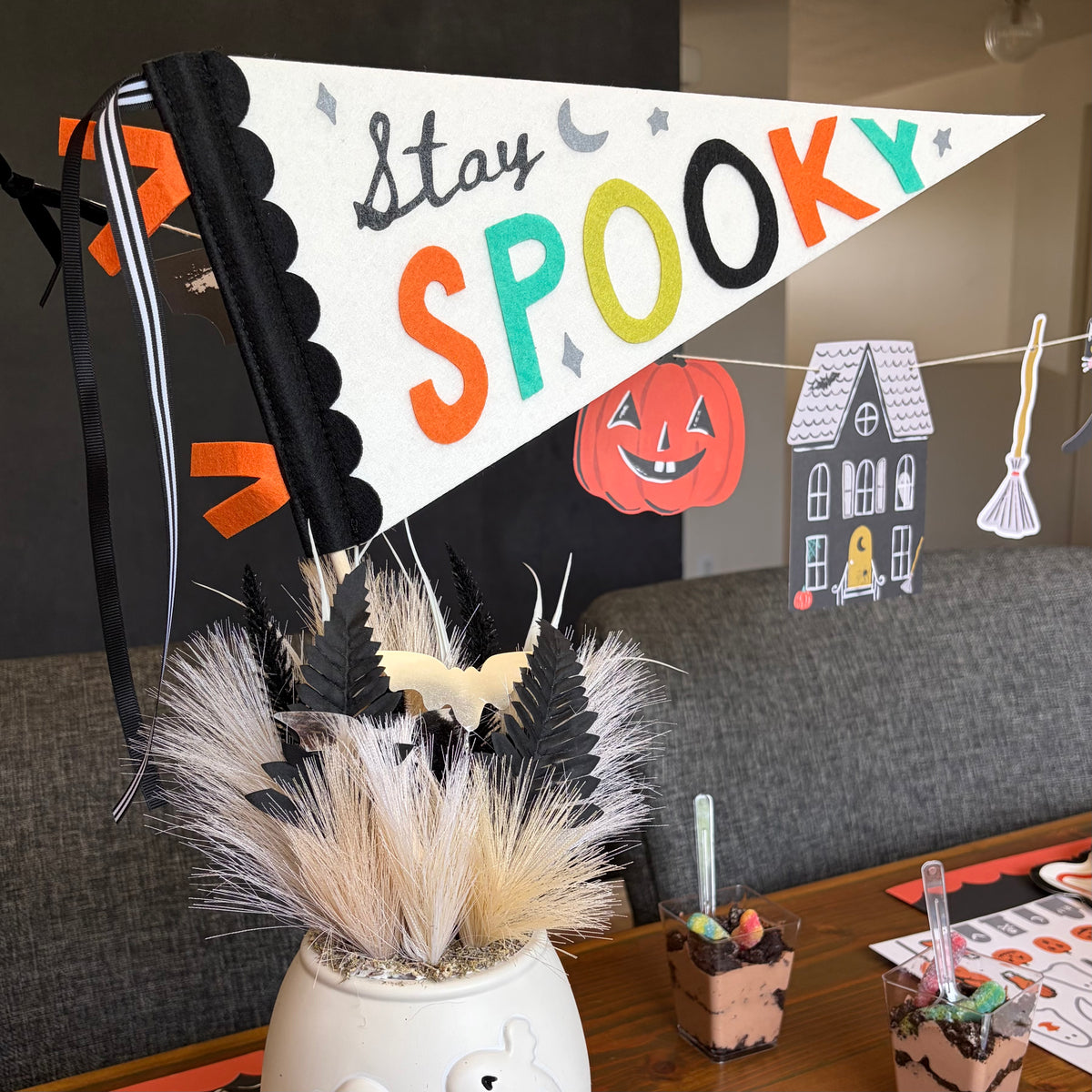 White Stay Spooky felt pennant flag with orange lettering and a pumpkin, propped in a vase with pampas grass beside black taper candles