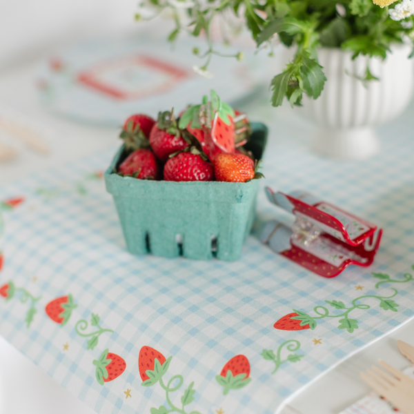 Strawberry gingham table runner styled on a table with a green basket of fresh strawberries, wildflowers, and strawberry-print party favors.