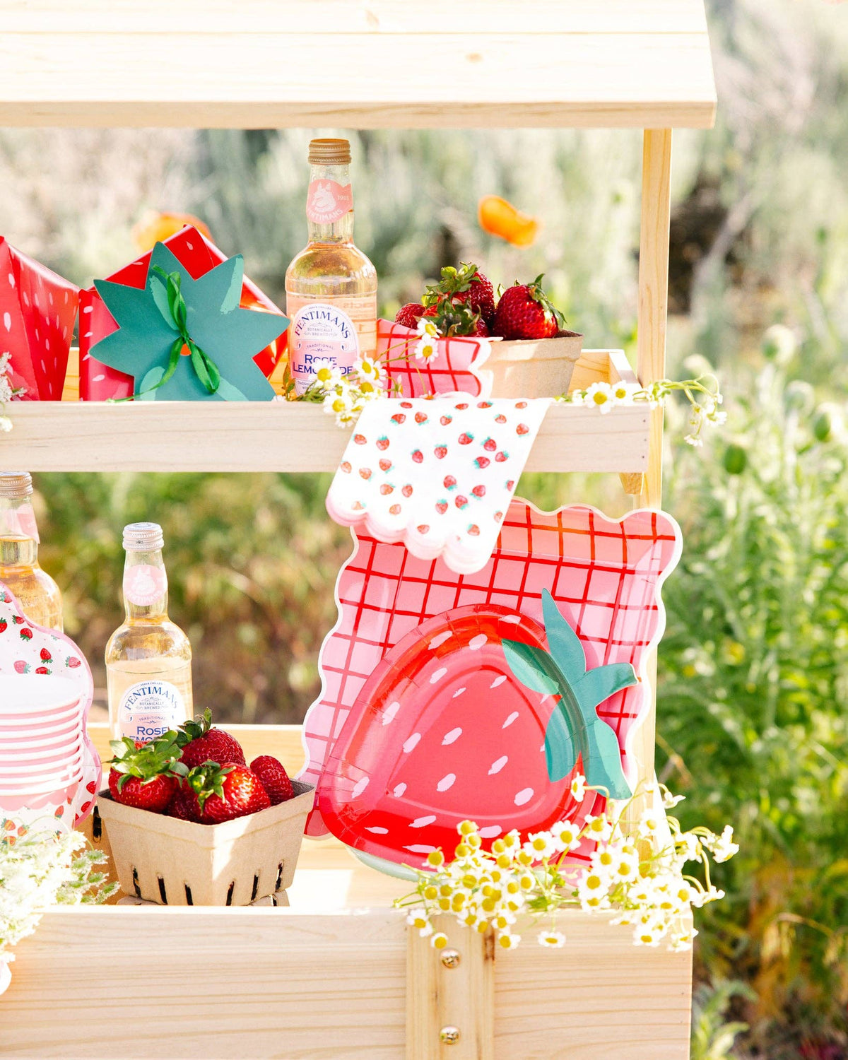 Wooden strawberry stand display with the full Strawberry Collection styled together — Strawberry Paper Guest Towels, strawberry-shaped paper plates, pink-and-red grid plaid paper plates, strawberry-shaped gift bags, fresh strawberries in paper berry baskets, daisies, and bottles of rose lemonade
