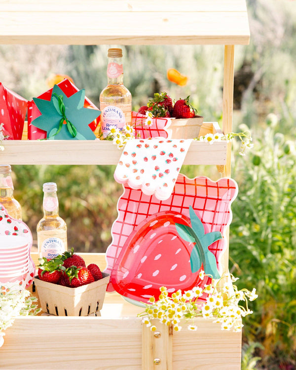 Wooden strawberry stand display with the full Strawberry Collection styled together — Strawberry Paper Guest Towels, strawberry-shaped paper plates, pink-and-red grid plaid paper plates, strawberry-shaped gift bags, fresh strawberries in paper berry baskets, daisies, and bottles of rose lemonade