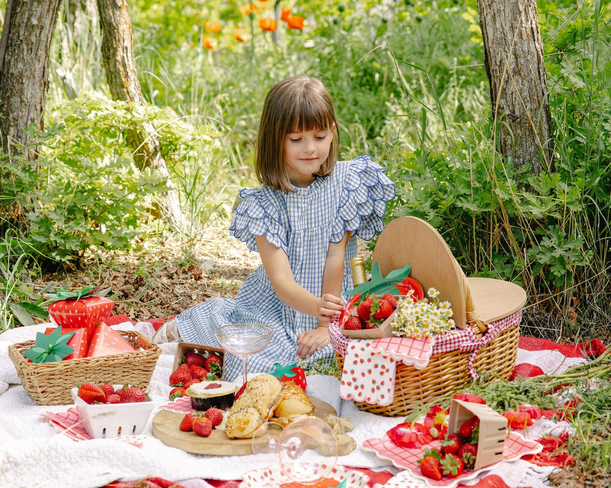 Outdoor summer strawberry picnic with a young girl in a blue gingham dress sitting on a white quilt and red gingham blanket beside a wicker picnic basket filled with fresh strawberries, surrounded by Strawberry Paper Guest Towels, strawberry-shaped gift bags, strawberry scalloped dinner plates, and a pink-and-red grid plaid plate