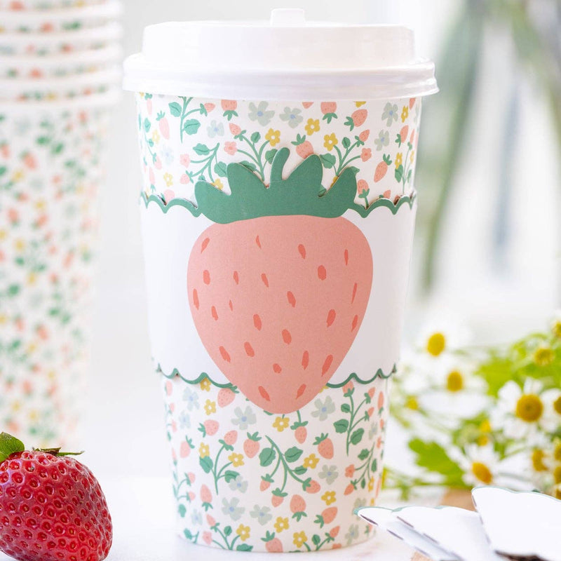 A 16 oz paper cup with a Cottagecore Chic strawberry motif sleeve sitting next to fresh strawberries.