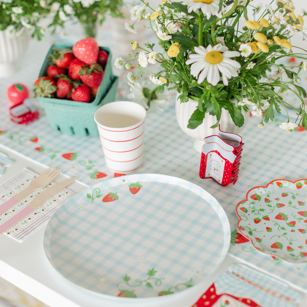 Berry first birthday flat-lay featuring the scalloped strawberry dessert plate, blue gingham strawberry dinner plate, strawberry preserves jar-shaped cocktail napkin, fresh strawberries in a green pint, and wildflowers