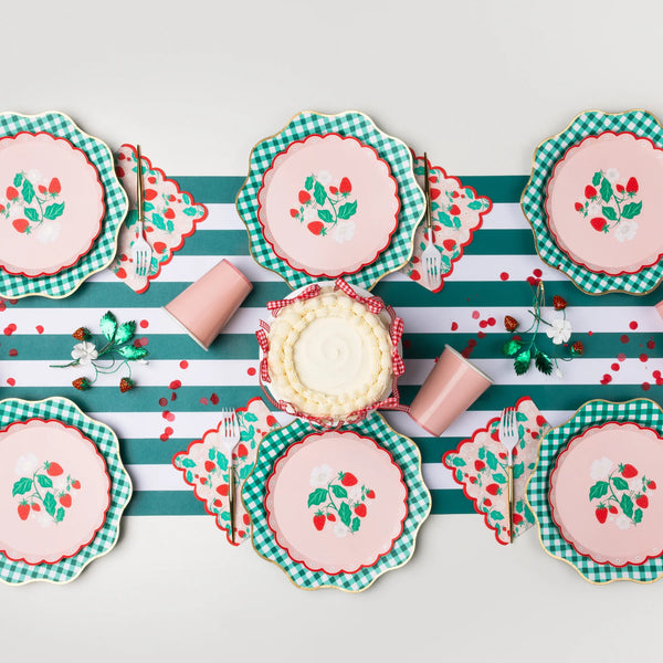 Strawberry scalloped paper cocktail napkins with red strawberries and white blossoms styled on a green and white striped runner with pink strawberry dessert plates, green gingham dinner plates, pink paper cups, and a buttercream cake with gingham ribbon.