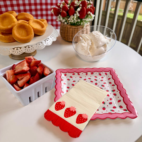 Strawberry shortcake bar setup with strawberry plates, strawberry napkins, whipped cream, and fresh strawberries.