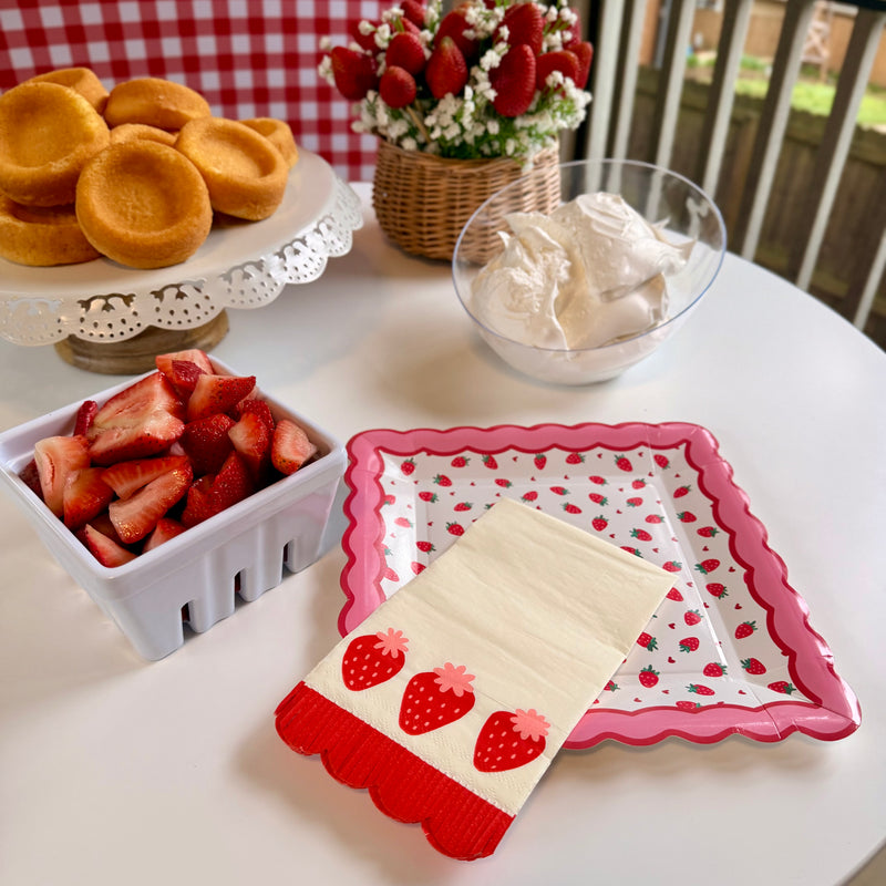 Strawberry shortcake bar setup with strawberry plates, strawberry napkins, whipped cream, and fresh strawberries.