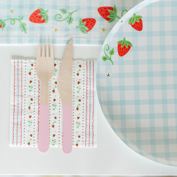 Close-up of a strawberry stripe paper cocktail napkin paired with the blue gingham strawberry-print dinner plate and pink-handled wooden cutlery on a strawberry-printed gingham table runner