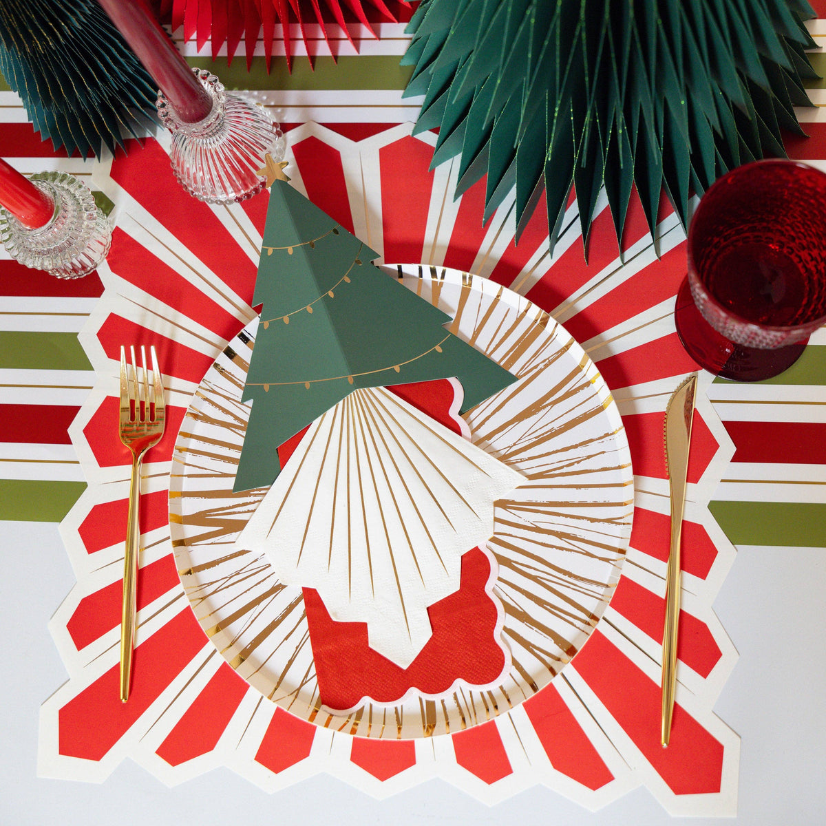 Overhead with a red-and-white striped starburst placemat, green paper fan rosettes, a green gnome figure, a red drink, and a gold fork.
