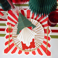 Overhead with a red-and-white striped starburst placemat, green paper fan rosettes, a green gnome figure, a red drink, and a gold fork.