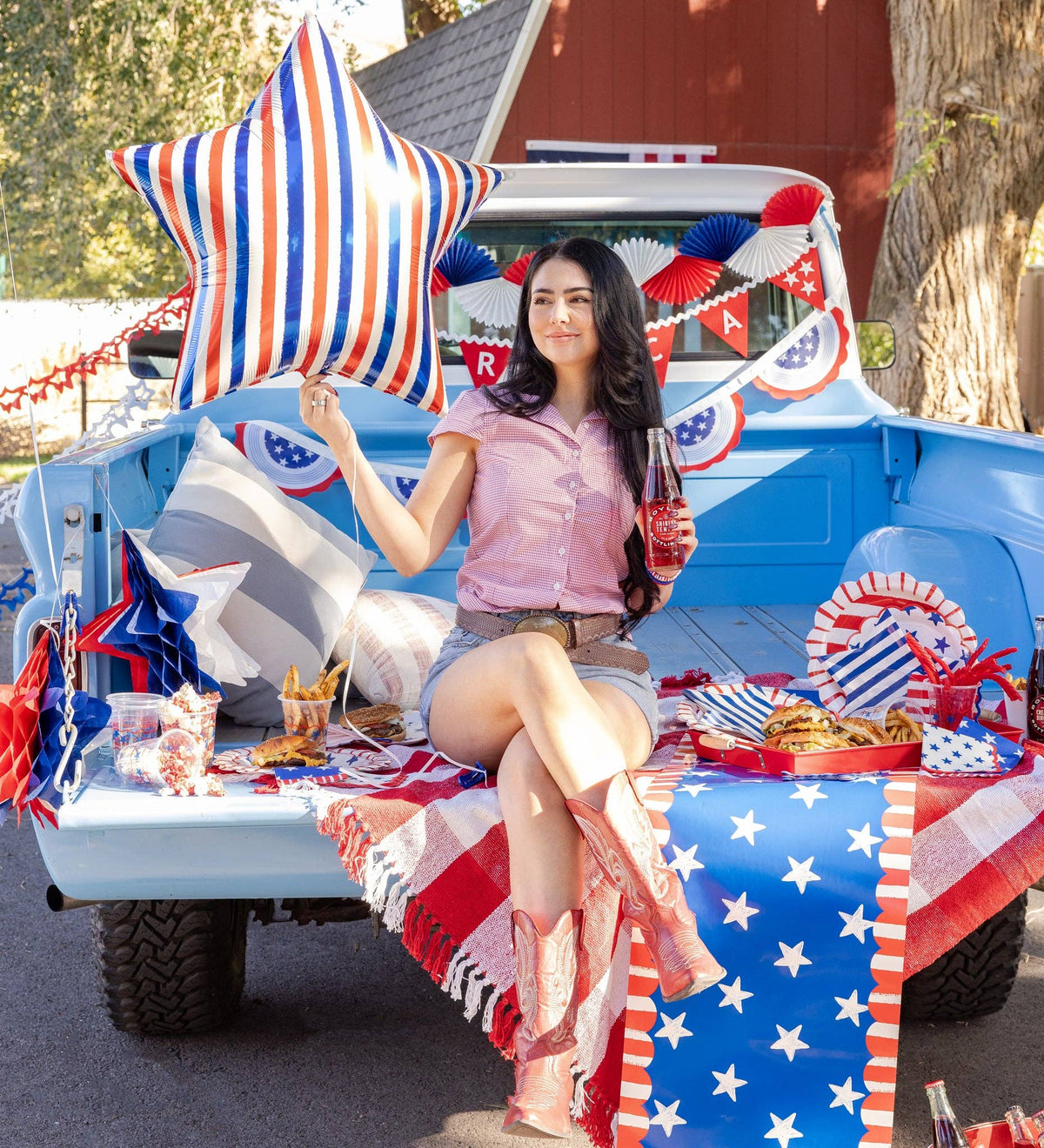 Woman sitting in vintage truck bed holding striped star mylar balloon at a patriotic outdoor picnic with American flag decor and party supplies
