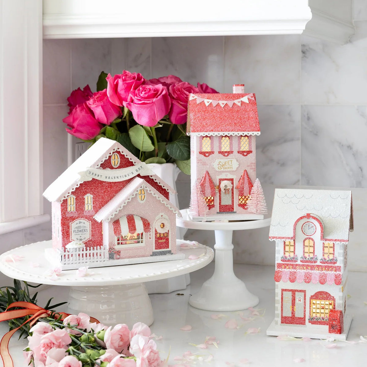 Display of three valentine gingerbread house pieces on white cake stands with pink flowers and valentines decorations.