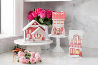 Lifestyle display of the three valentine gingerbread house pieces on white cake stands with pink roses.