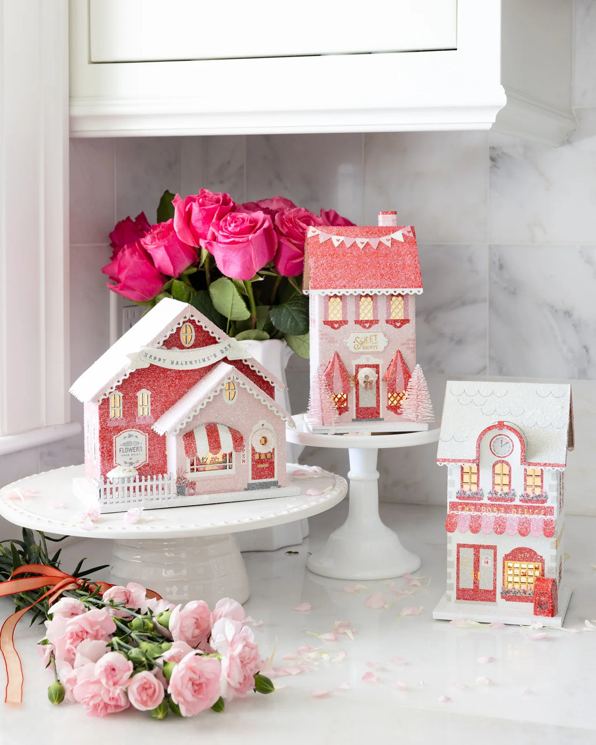 Full set of the three valentine village putz house designs illuminated on a marble counter with pink flowers.