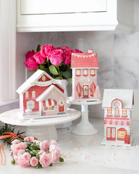 Full set of the three valentine village putz house designs illuminated on a marble counter with pink flowers.