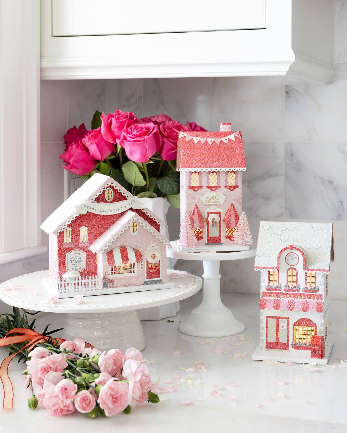 Full set of the valentine village houses displayed on a counter, with the sweet shoppe decor glowing and surrounded by pink roses.
