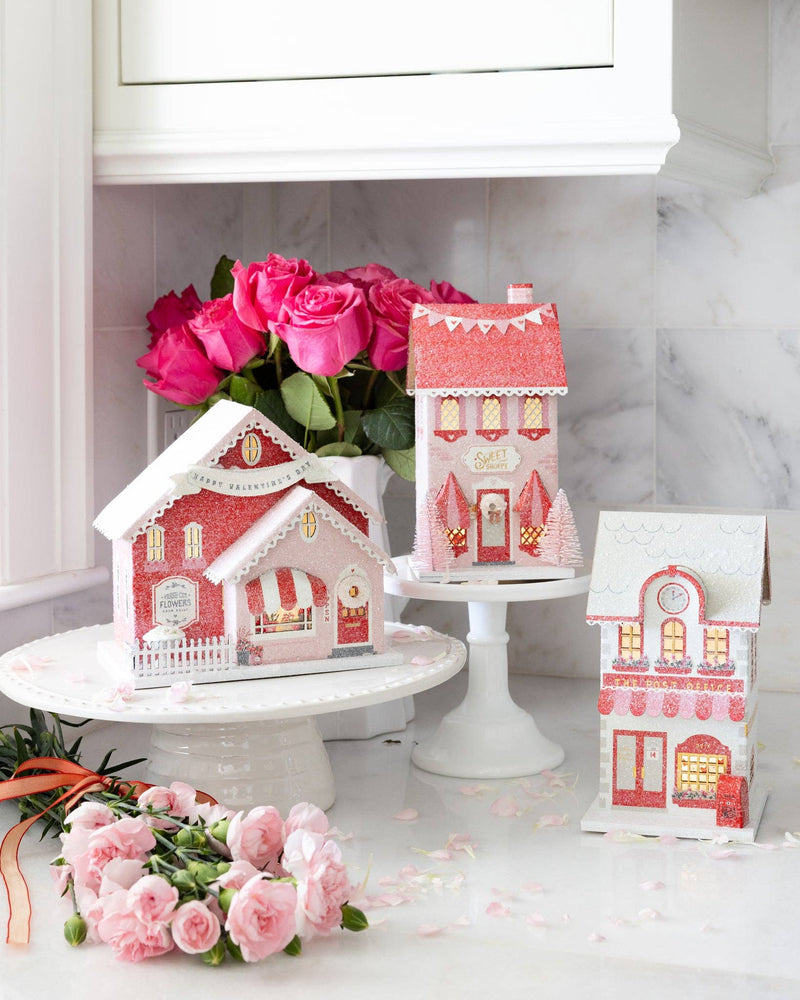 Full set of the valentine village houses displayed on a counter, with the sweet shoppe decor glowing and surrounded by pink roses.