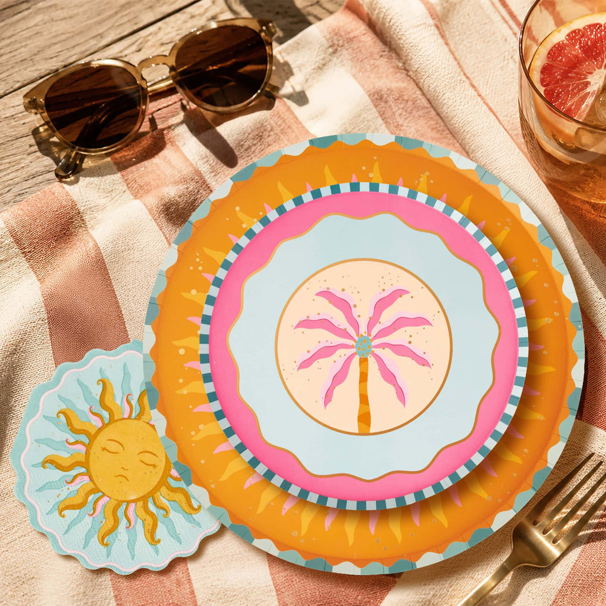 Boho summer place setting with a sun shaped napkin, layered sunburst and palm-print plates, and sunglasses on a striped towel.