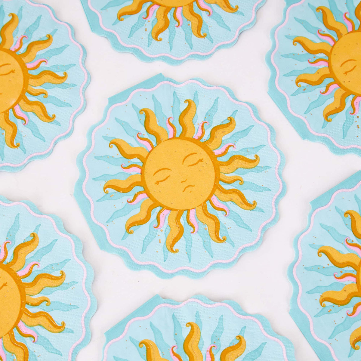 Sun shaped paper cocktail napkins scattered on white, showing the celestial sun face and wavy pink-trimmed edge.