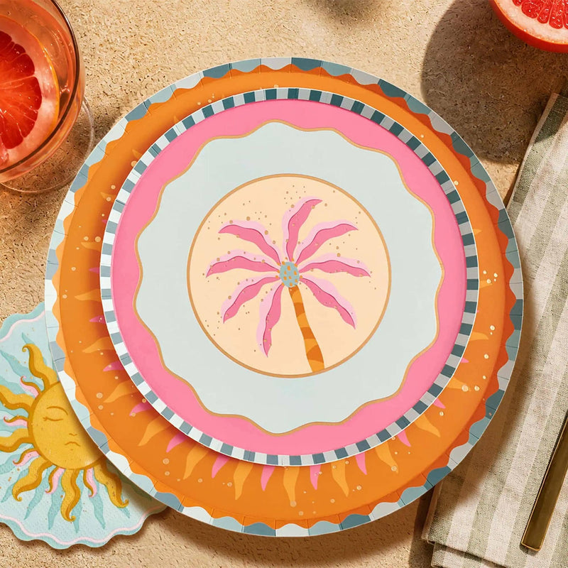 Sunburst dinner plate layered under a pink palm plate with orange cocktails and olives on a sunny table.