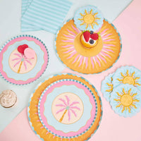 Orange sunburst dinner plate styled with sun-shaped napkins, a pink palm plate, and striped napkins in a pastel flatlay with a berry cupcake.