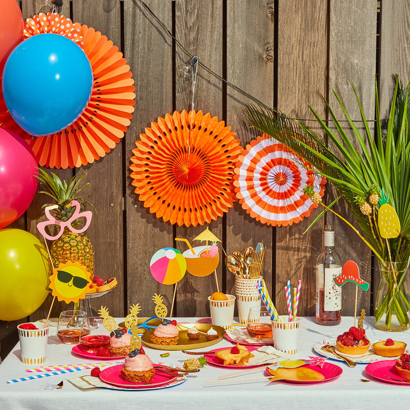 A full Modern Luau Table Backdrop with Tropical Party Fans hung on a wooden fence next to balloons and tropical treats.