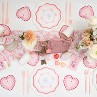 Overhead view of coquette Valentine's Day party table with swan love birds scalloped plates, heart shaped napkins, pink cutlery and floral table runner with roses and carnations