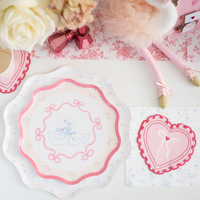 Close-up of swan love birds scalloped dinner plate with heart shaped napkin and pink bow coquette table setting with white roses and pink metallic accents