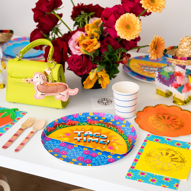 This photo shows a colorful taco-themed party table with taco plates, flowers, fiesta decorations, and party tableware.